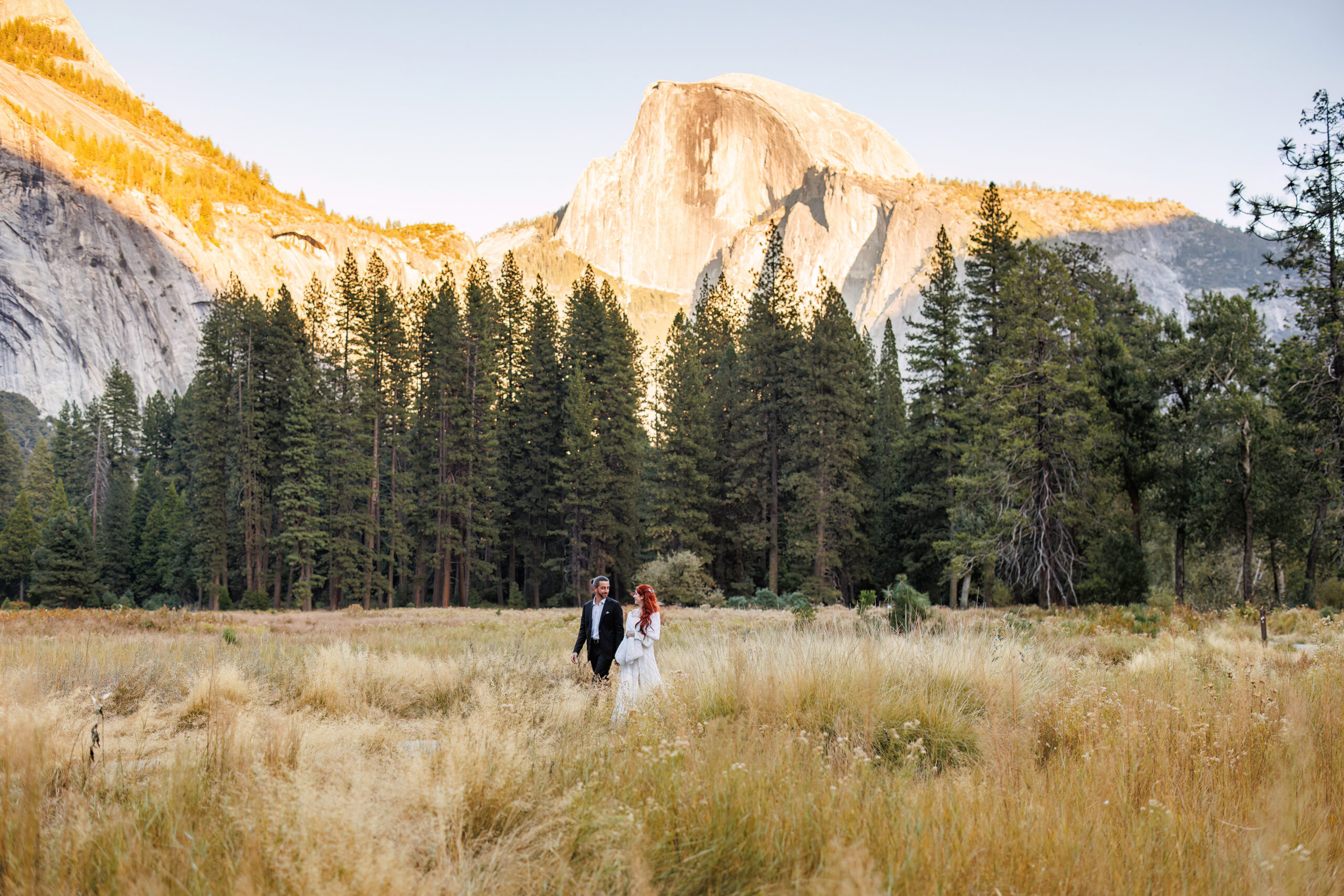 a couple walking through a meadow in yosemite with mountains in the background 